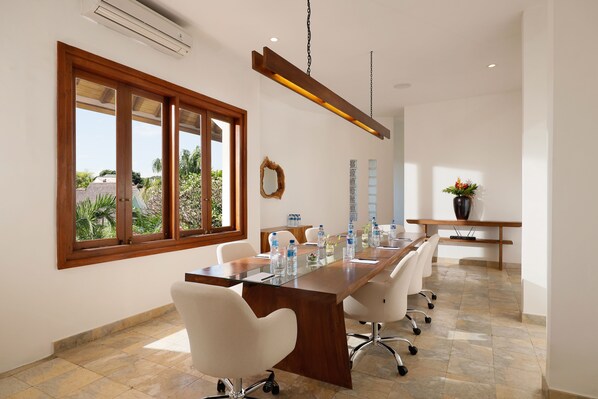 A conference room with a long wooden table, white chairs, a large window, and a hanging light fixture. - Hotel Puri Tempo Doeloe (Denpasar)