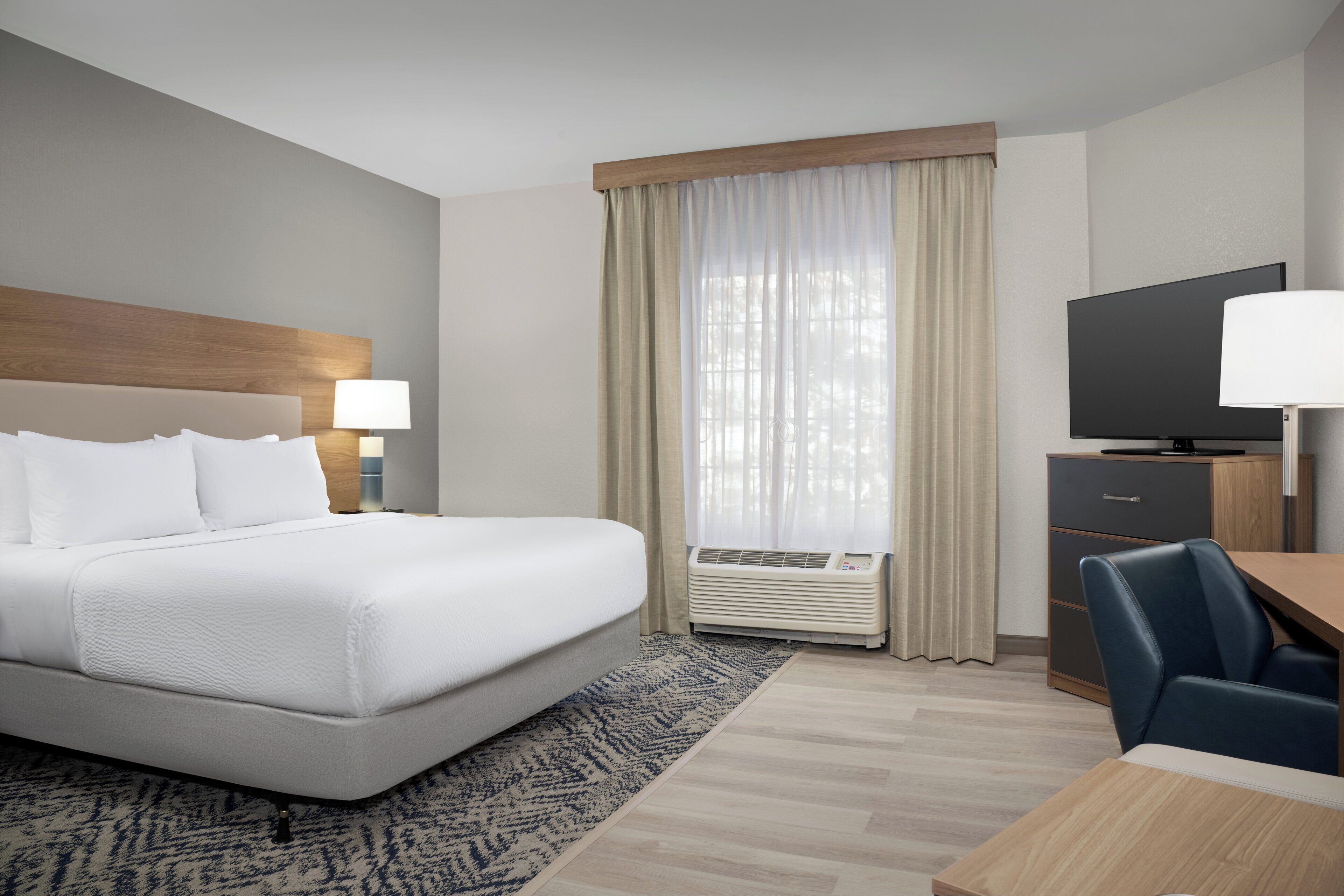 A hotel room with a bed, a desk, a chair, a television, and an air conditioning unit.