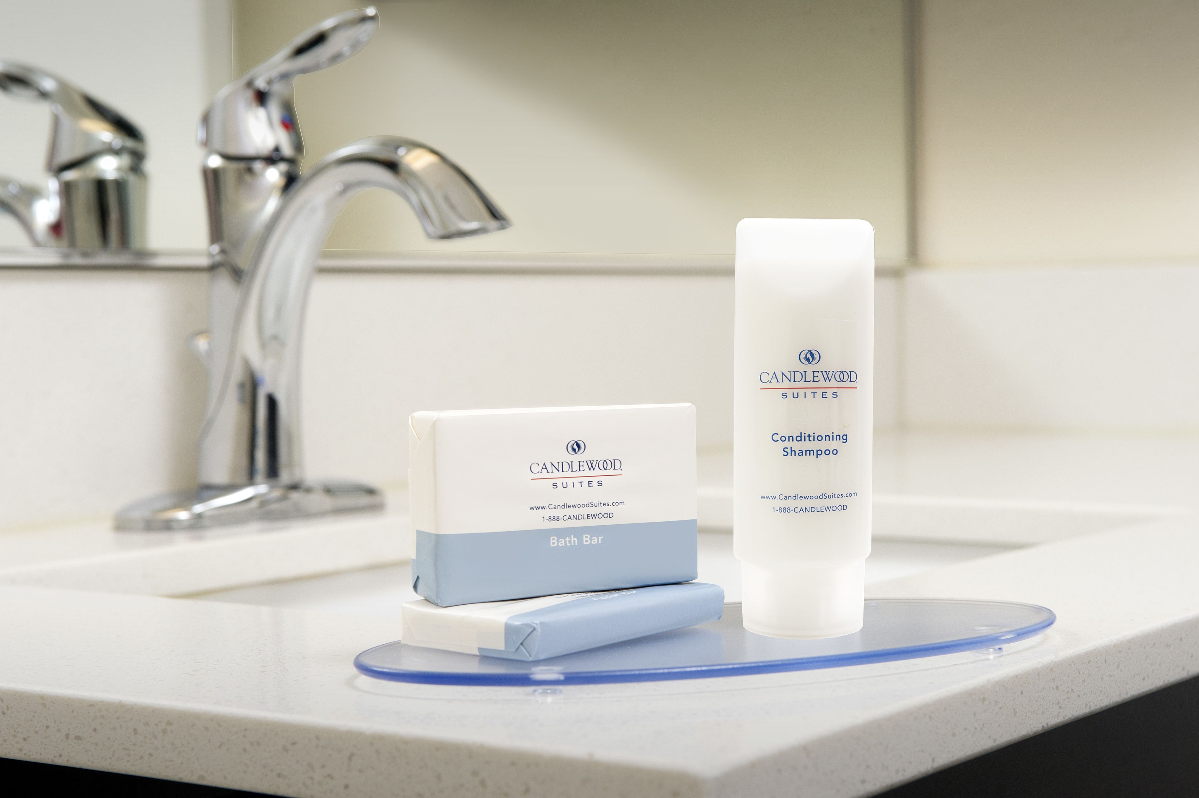 A bathroom sink with a white countertop, a blue soap dish, a white bottle of Candlewood Suites conditioning shampoo, and a white soap bar.