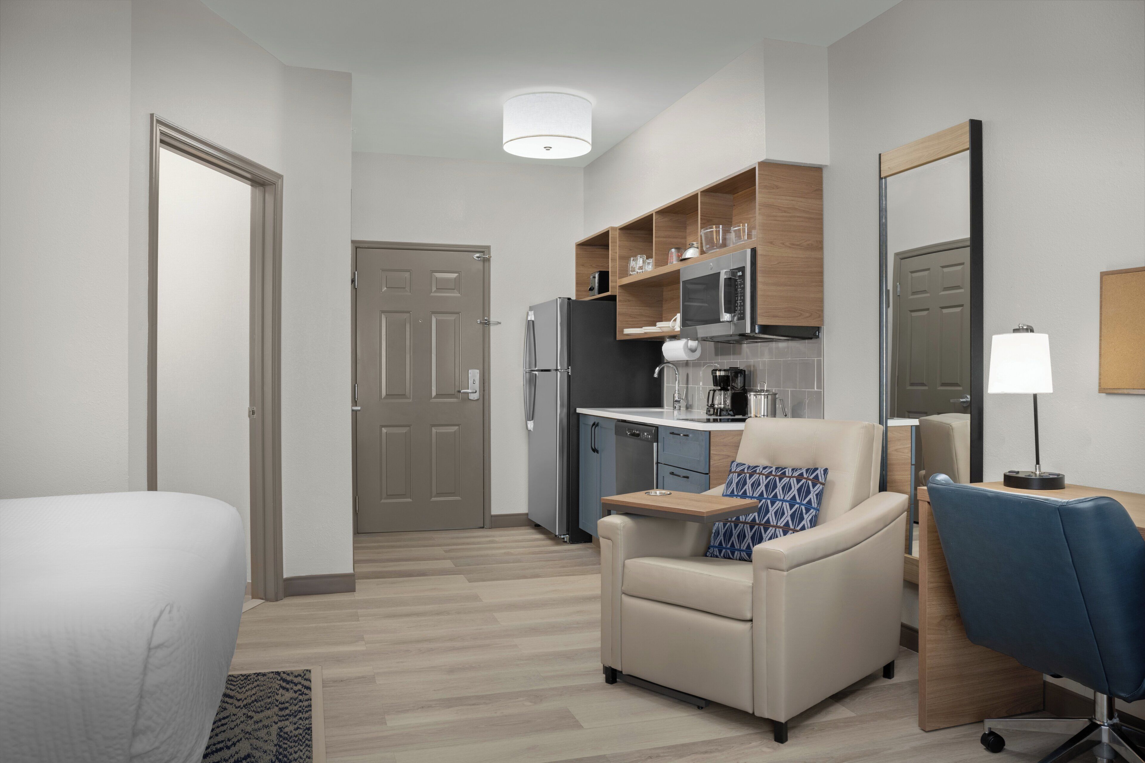 A compact hotel room with a bed, a small seating area with a chair and a table, a kitchenette with a refrigerator and microwave, and a door leading to another room.