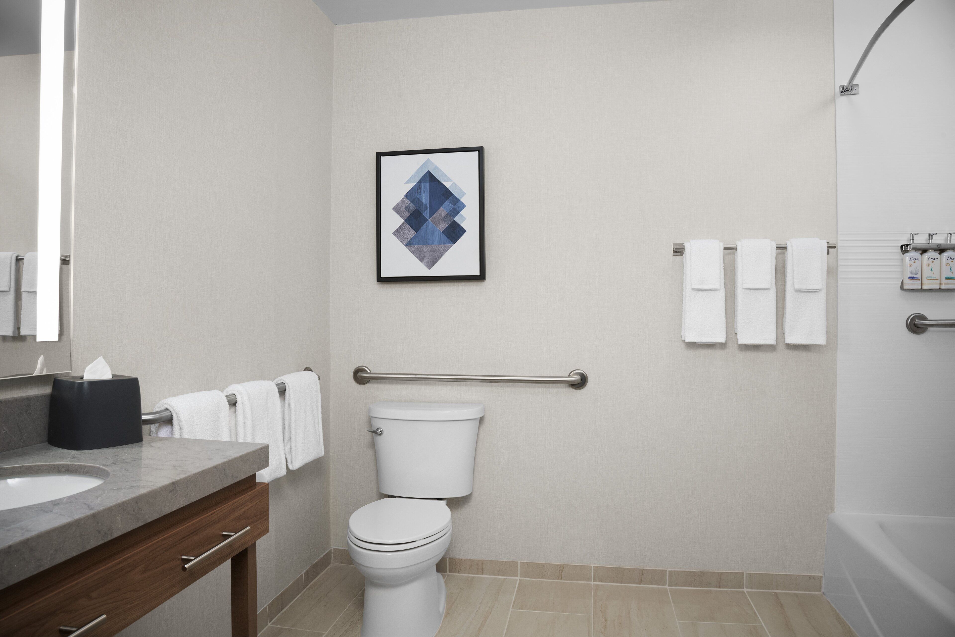 A bathroom with a toilet, a hand dryer, a towel rack with towels, a framed picture on the wall, and a hand sanitizer dispenser.