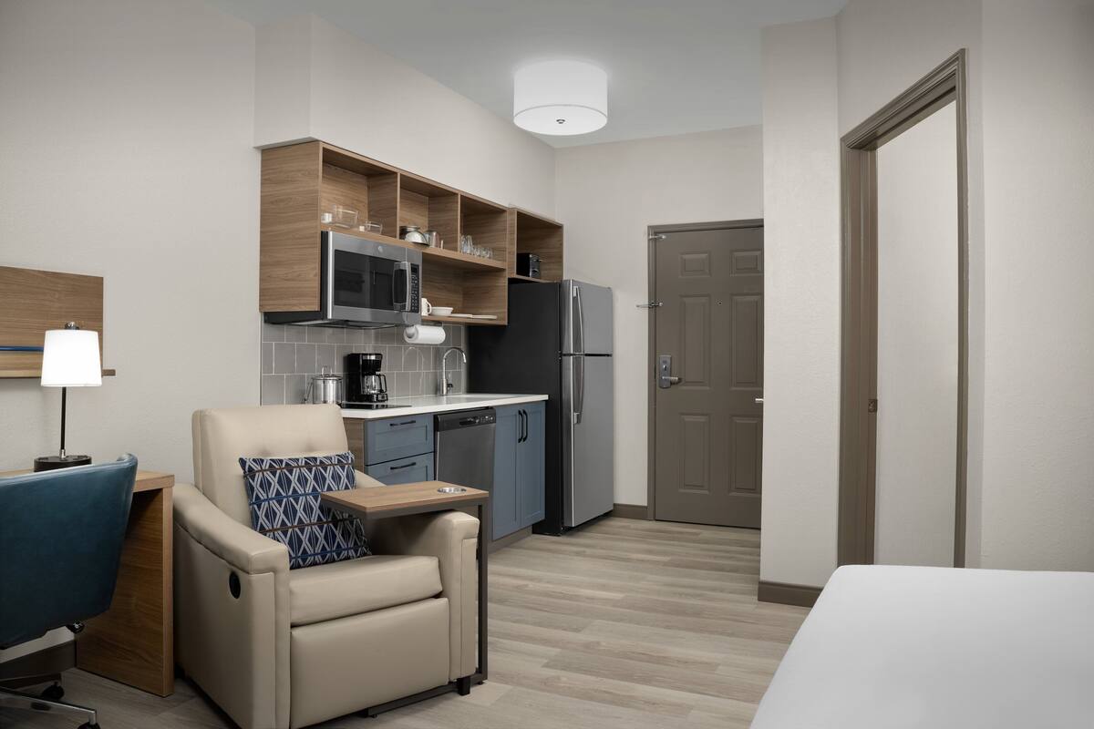 A compact hotel room with a kitchenette, a sofa with a pillow, a desk with a lamp, and a door.