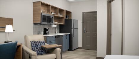 Habitación de hotel compacta con kitchenette, un sofá con un cojín, un escritorio con lámpara y una puerta.