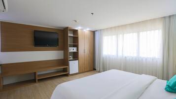 A hotel room with a bed, a TV mounted on the wall, a small kitchenette, and a window with curtains.