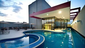 Una piscina moderna al aire libre con diseño curvo, rodeada de sillones de descanso y sombrillas, y adyacente a un edificio con grandes ventanales.