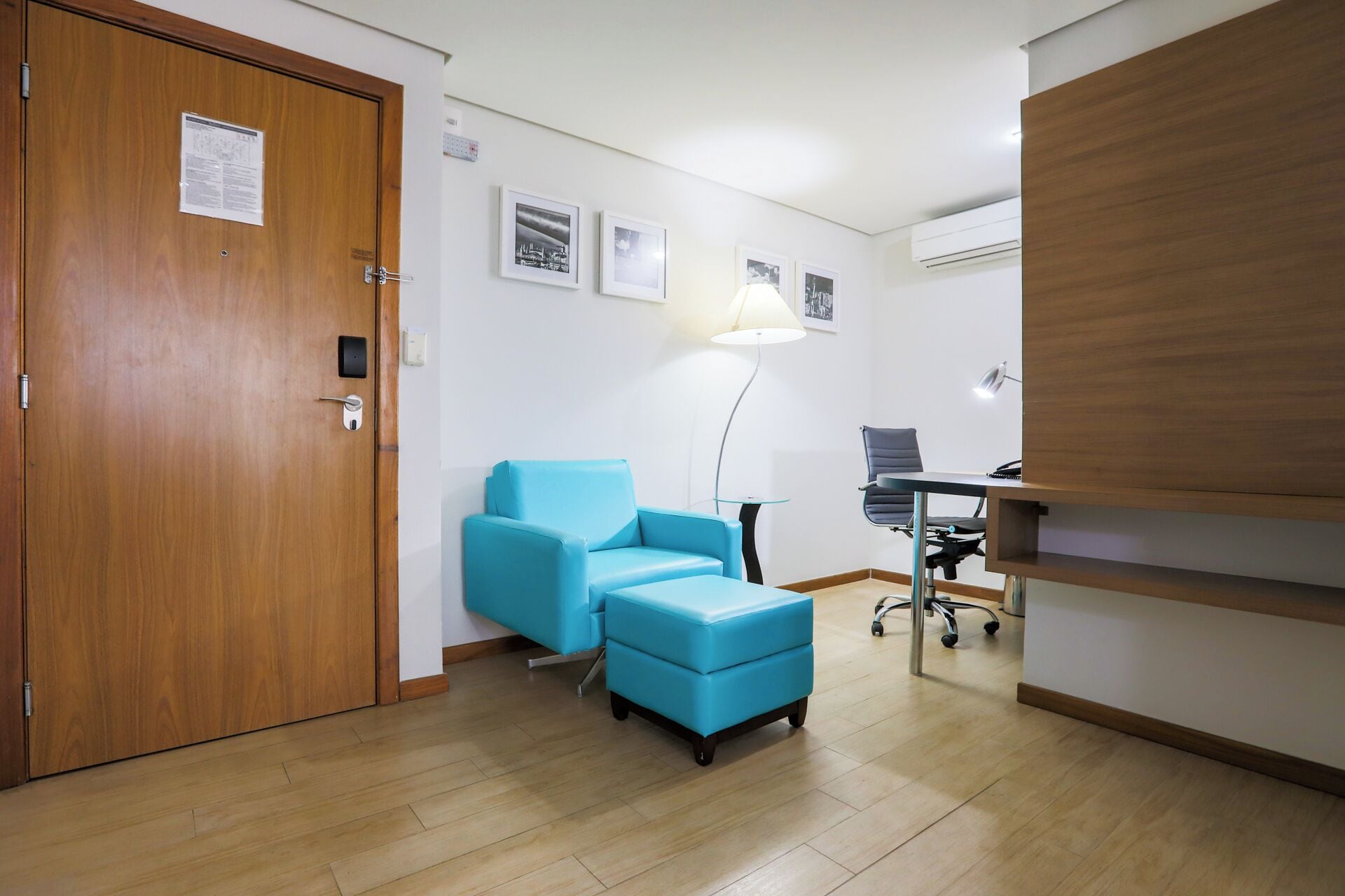 A room with a wooden door, a blue sofa, a desk with a chair, and a lamp.