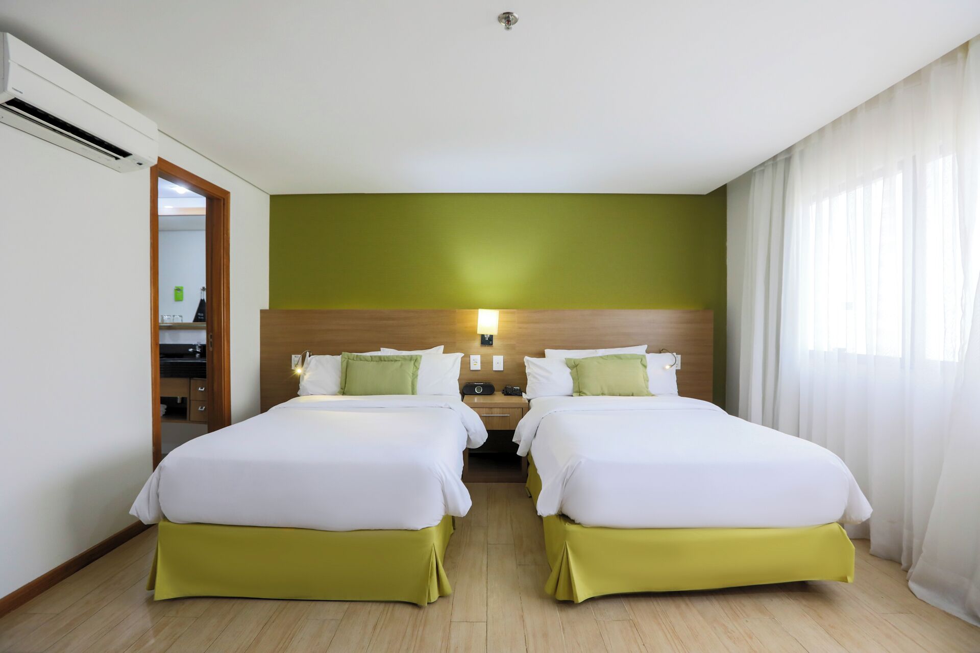 A hotel room with two beds, a wooden headboard, a nightstand with a lamp, and a bathroom visible through an open door.
