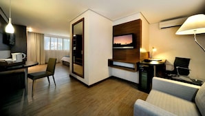 Una habitación de hotel moderna con una televisión de pantalla plana montada en un panel de madera, un escritorio con una silla, un sofá y una mesa de comedor con una silla.