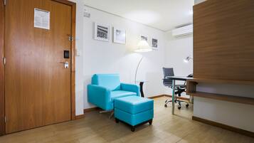A room with a wooden door, a blue sofa, a small table, a lamp, and a desk with a chair.