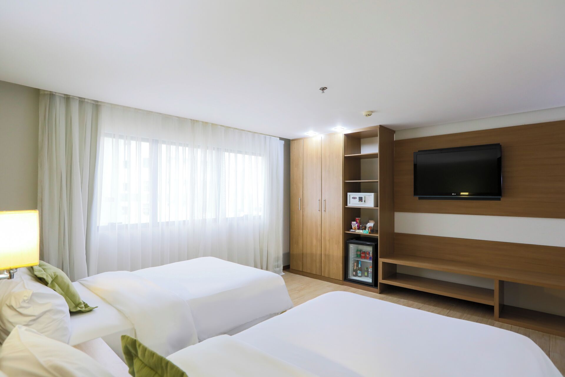 A hotel room with two beds, a TV mounted on the wall, a minibar, and a large window with curtains.