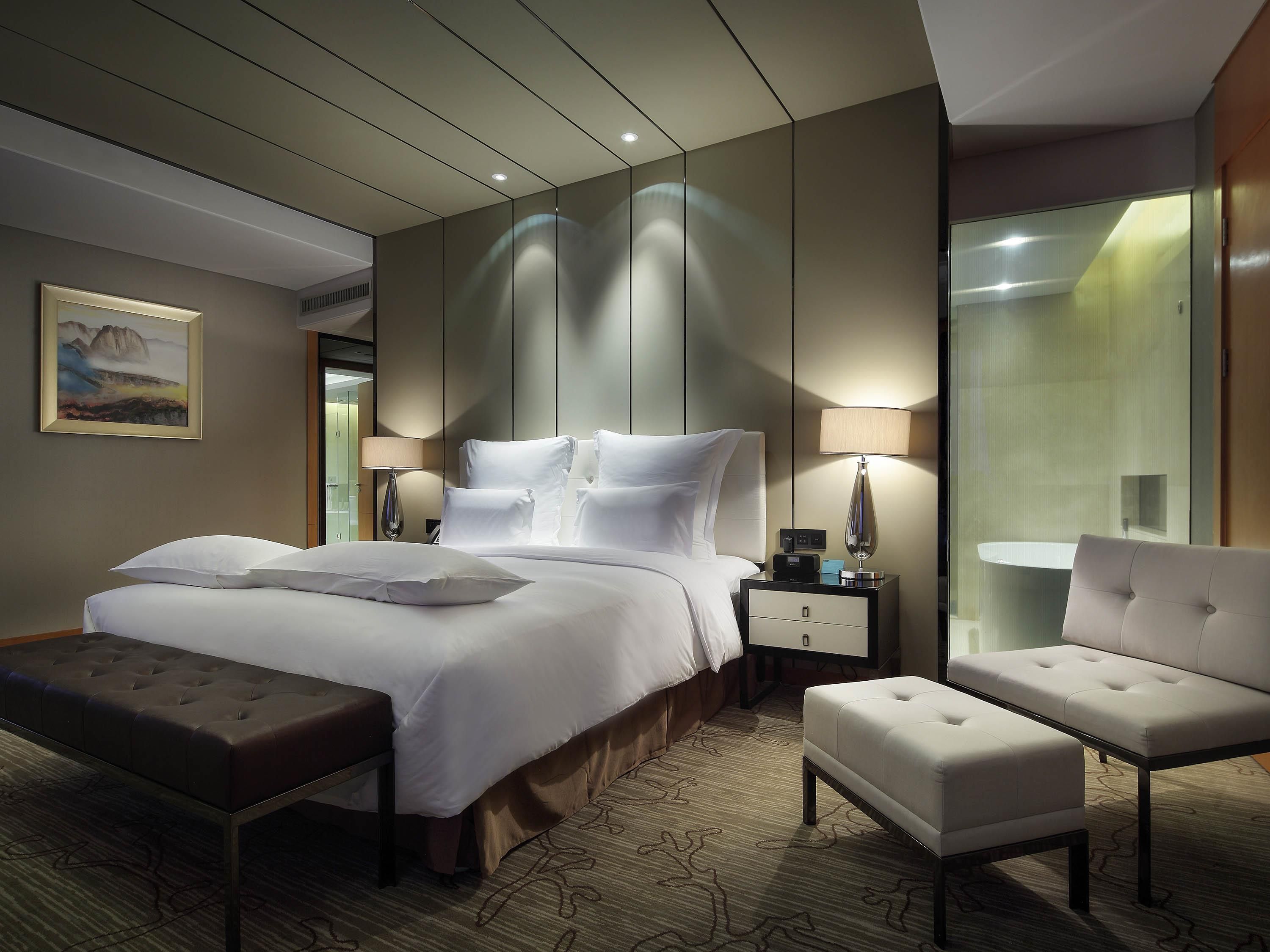 A modern hotel room with a large bed, a bedside table with a lamp, a bench, and a chair.