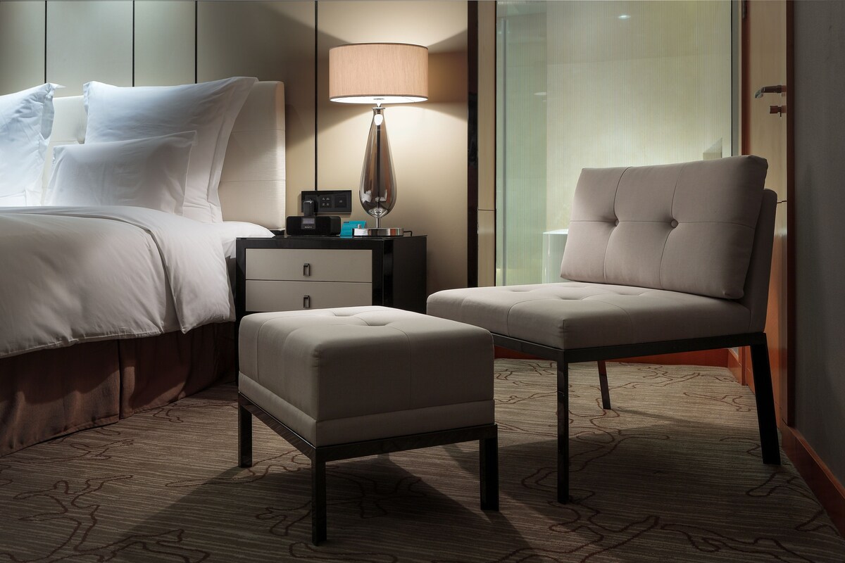 Una habitación de hotel con una cama, una mesita de noche, una lámpara y dos otomanas.
