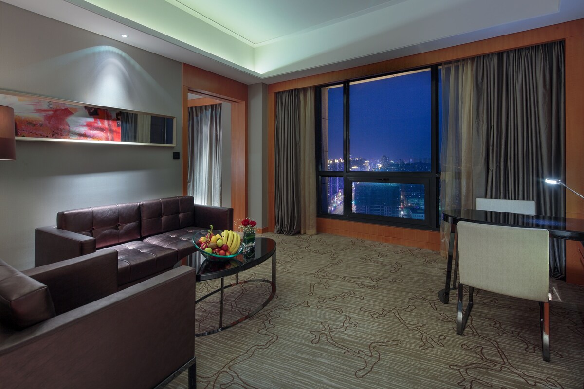 Habitación de hotel moderna con sofá de cuero, una mesa de comedor y un gran ventanal con vista a la ciudad de noche.