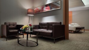 A modern hotel room with a brown leather sofa, a glass coffee table with fruits and flowers, a bedside table, and a large window with a skylight. - Pullman Anshan Time Square (Anshan)