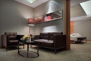 Una habitación de hotel moderna con un sofá de cuero marrón, una mesa de centro de vidrio con frutas y flores, una mesita de noche y un ventanal con tragaluz.