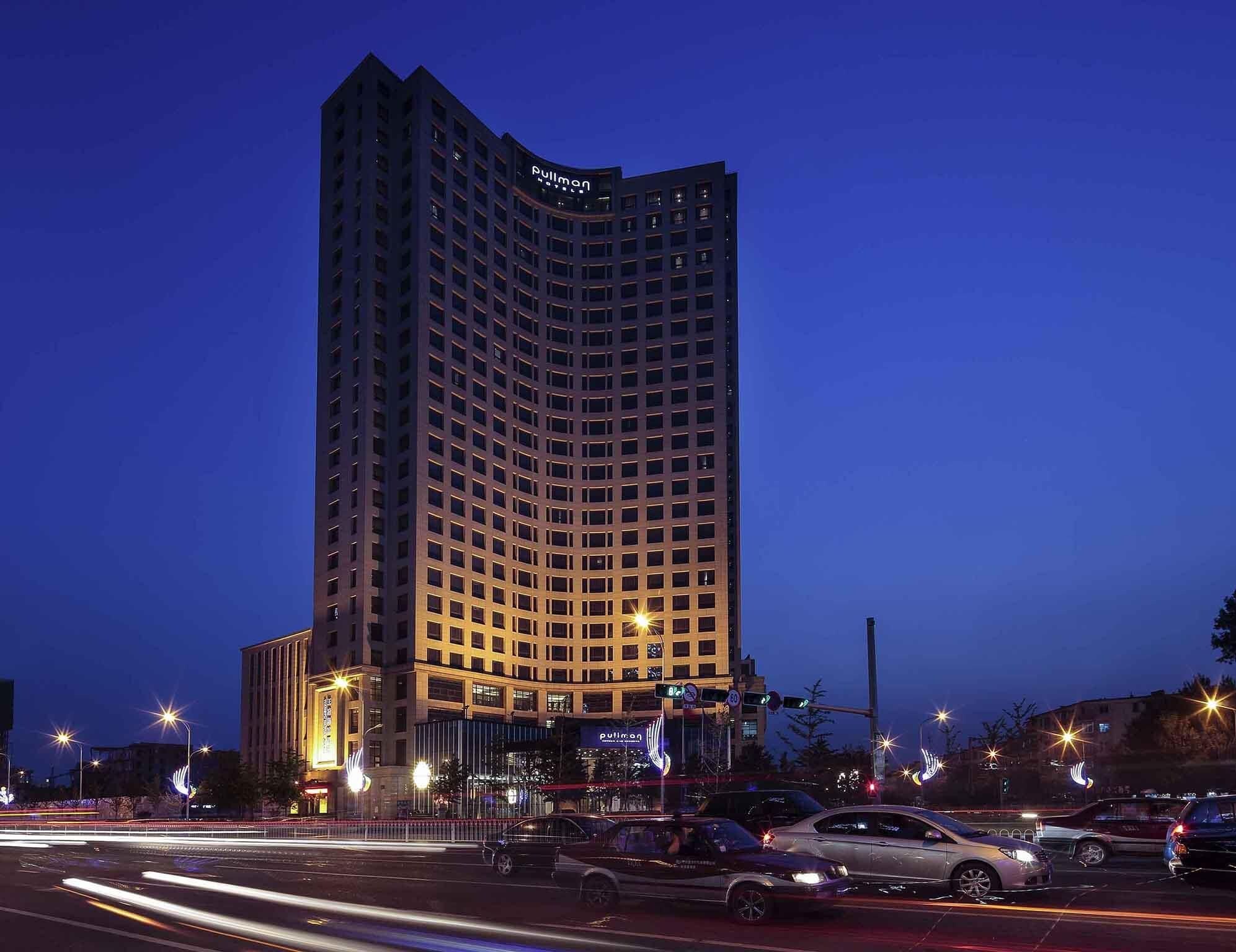 A high-rise hotel with a curved facade, illuminated windows, and a prominent 'Pullman' sign at the top.