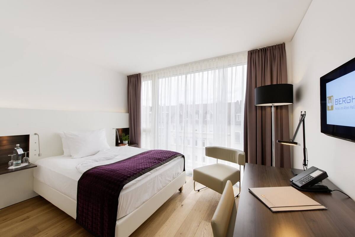 Habitación de hotel con cama, escritorio, silla, televisión y ventana con cortinas.