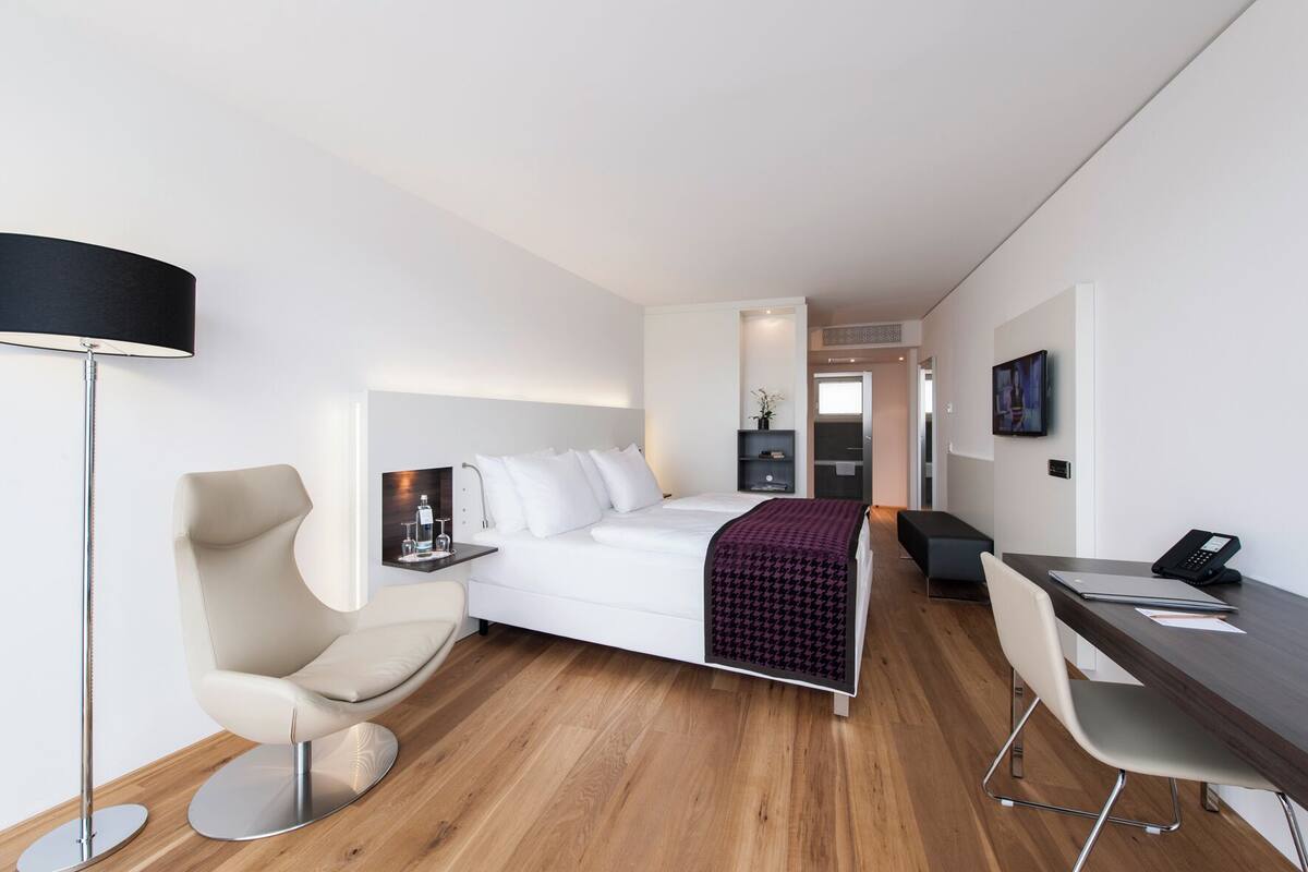 Una habitación de hotel moderna con una cama, un escritorio, una silla, una lámpara de pie y una pequeña mesa con una botella y vasos.