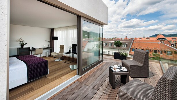 A modern hotel room with a balcony, a bed, a desk, a chair, and a TV.