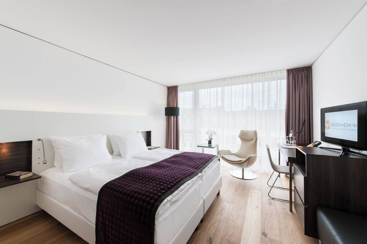 Una habitación de hotel moderna con una cama grande, un escritorio con computadora, una silla y un televisor.