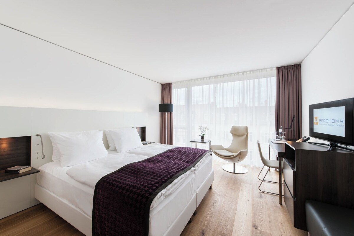 A modern hotel room with a large bed, a desk with a computer, a chair, and a television.