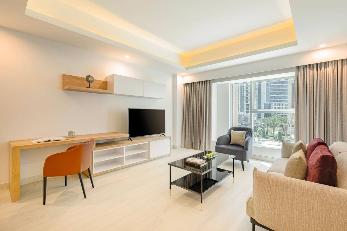 A modern living room with a flat-screen TV, a wooden desk, a comfortable sofa, and a large window with curtains.