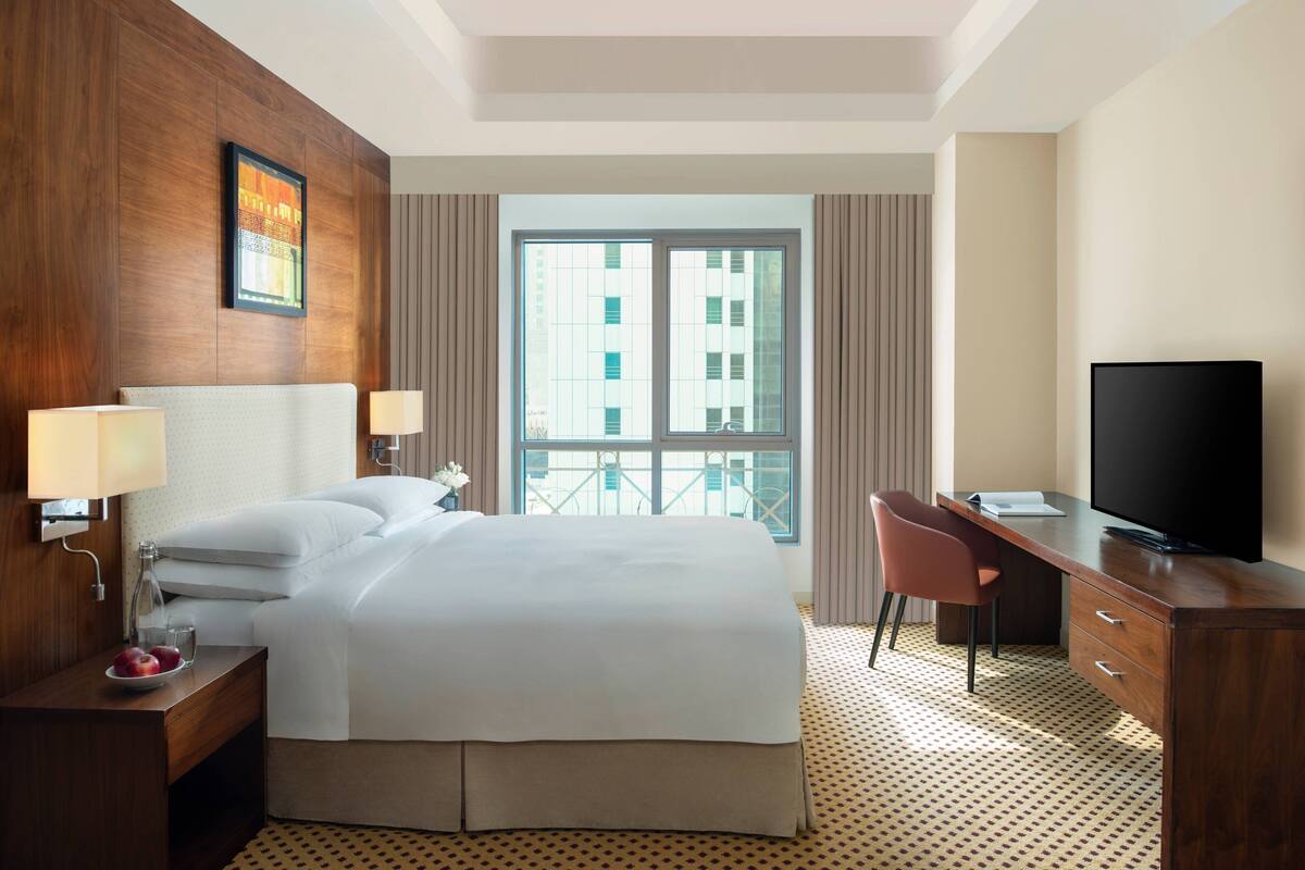 A hotel room with a large bed, a desk with a TV, a chair, and a window with a view of buildings.