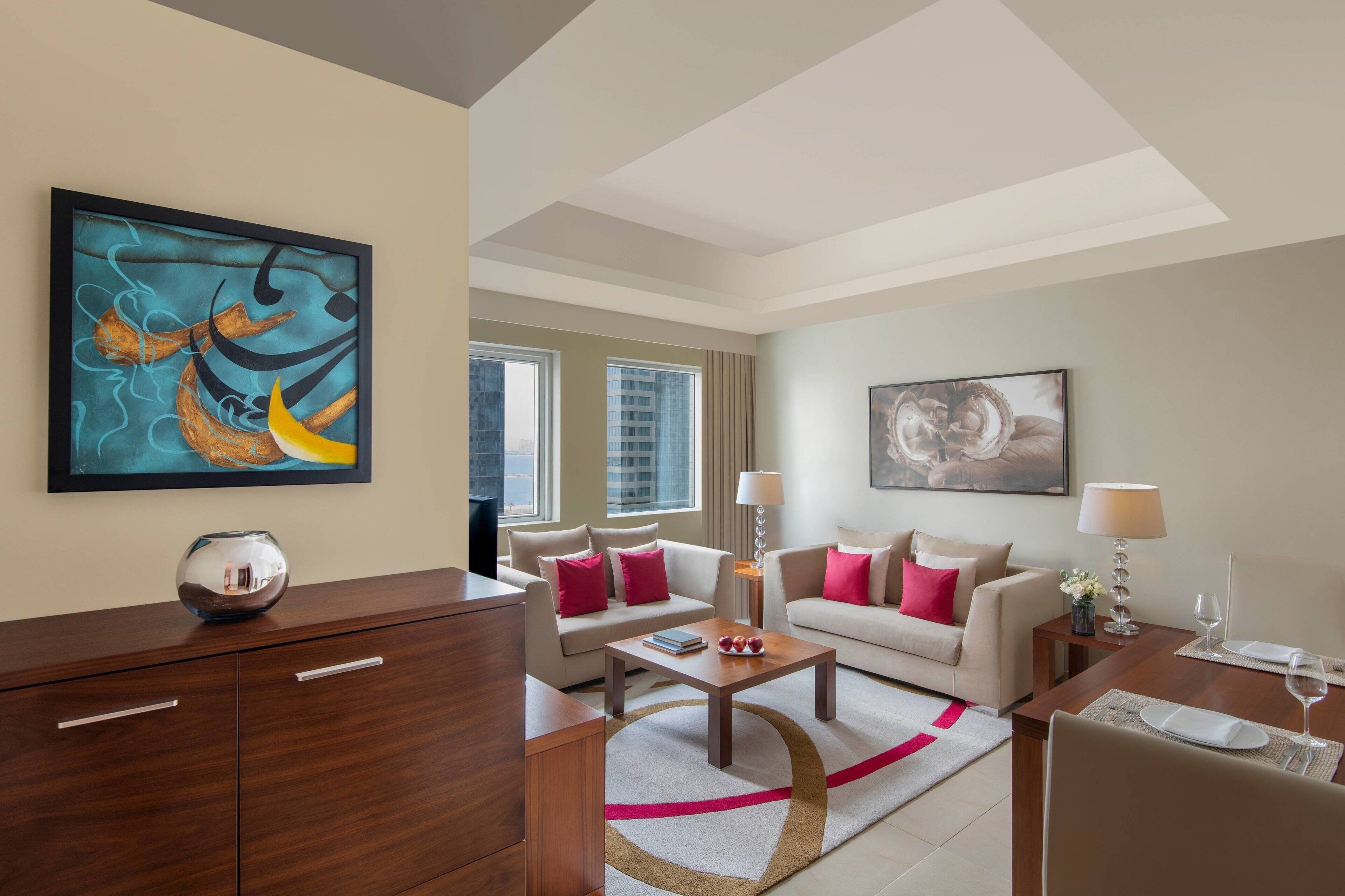 A modern hotel room with a living area featuring a sofa set, a coffee table, and a dining table with chairs. There is artwork on the walls and a view of the city through the windows.