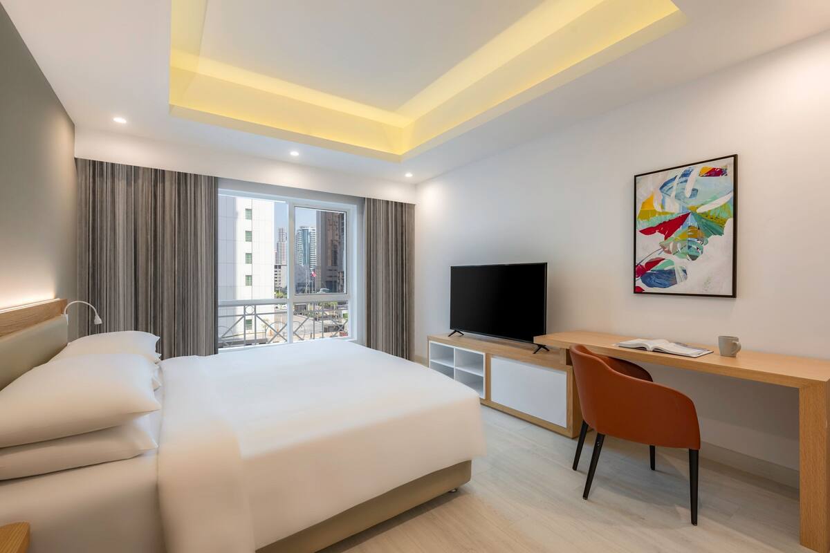 A modern hotel room with a large bed, a desk with a chair, a flat-screen TV, and a window with a city view.