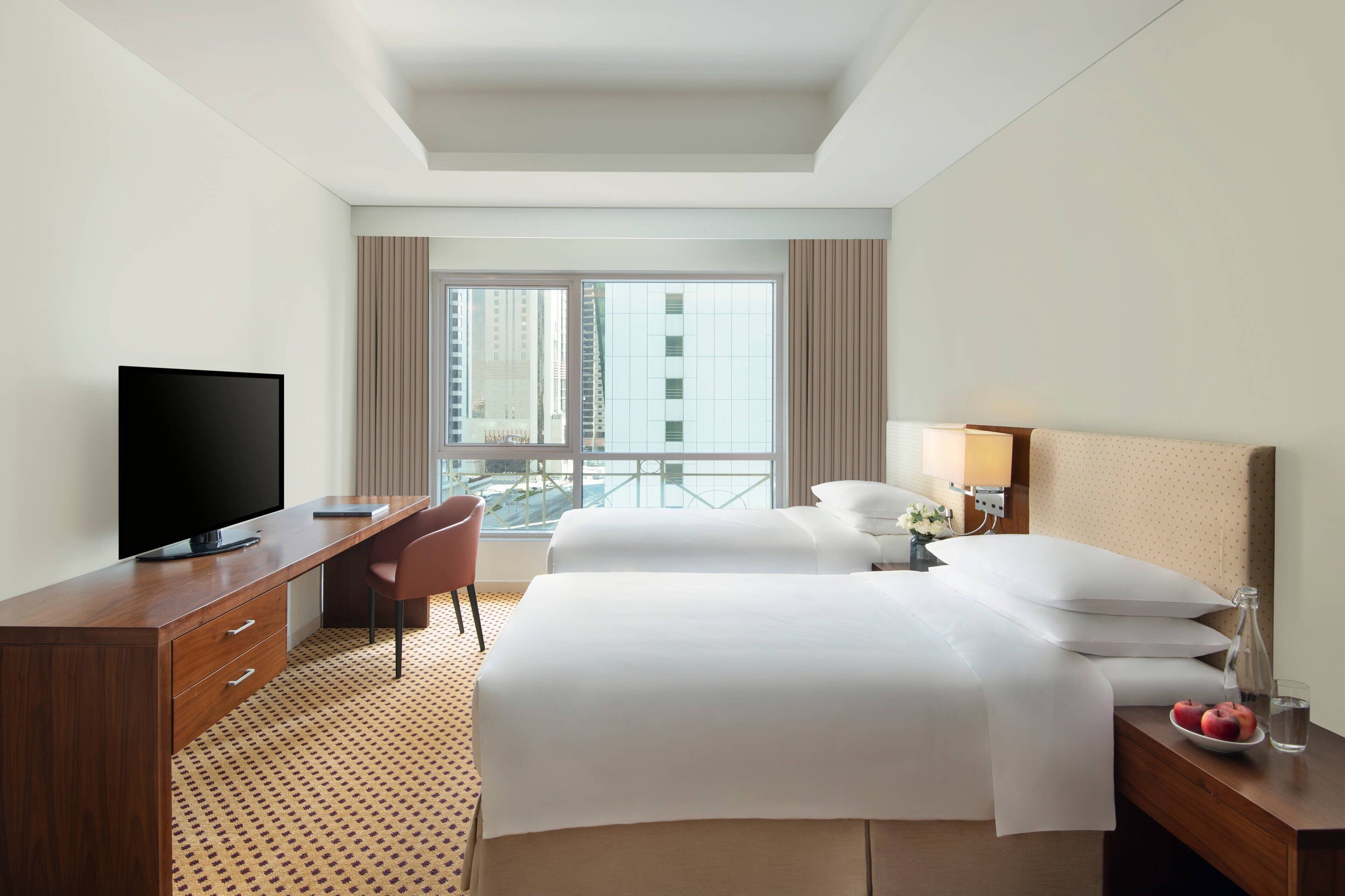 Une chambre d’hôtel comprenant un lit, un bureau avec une télévision, une chaise, une table de chevet avec une lampe et une fenêtre donnant sur des immeubles.