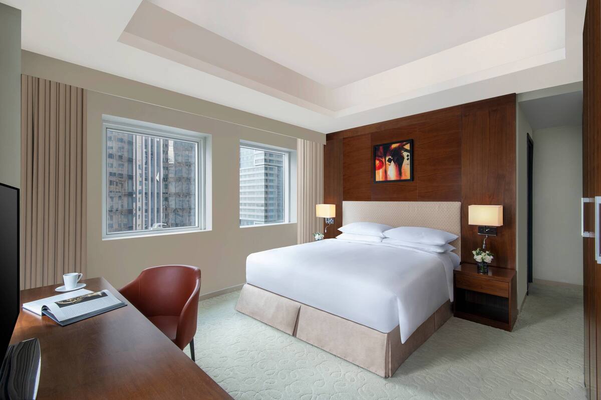 A hotel room with a large bed, two bedside lamps, a desk with a chair, and a window with blinds.