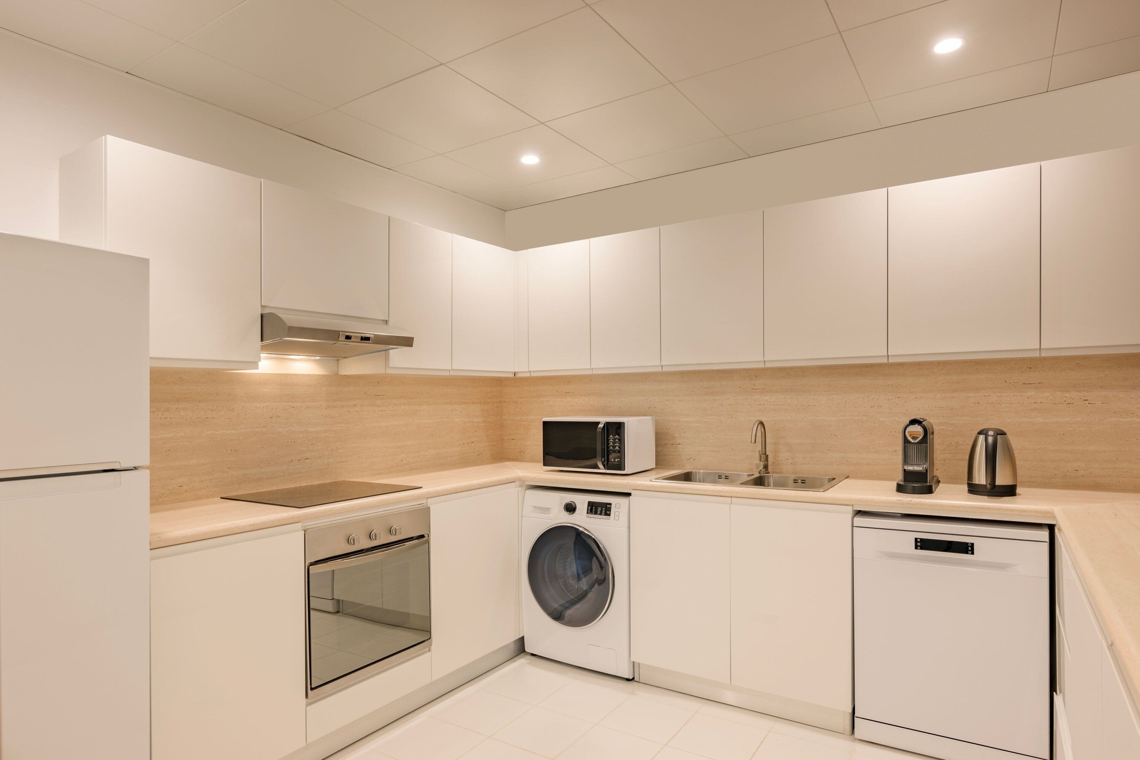 Una cocina moderna con armarios blancos, horno integrado, lavadora, microondas y cafetera.