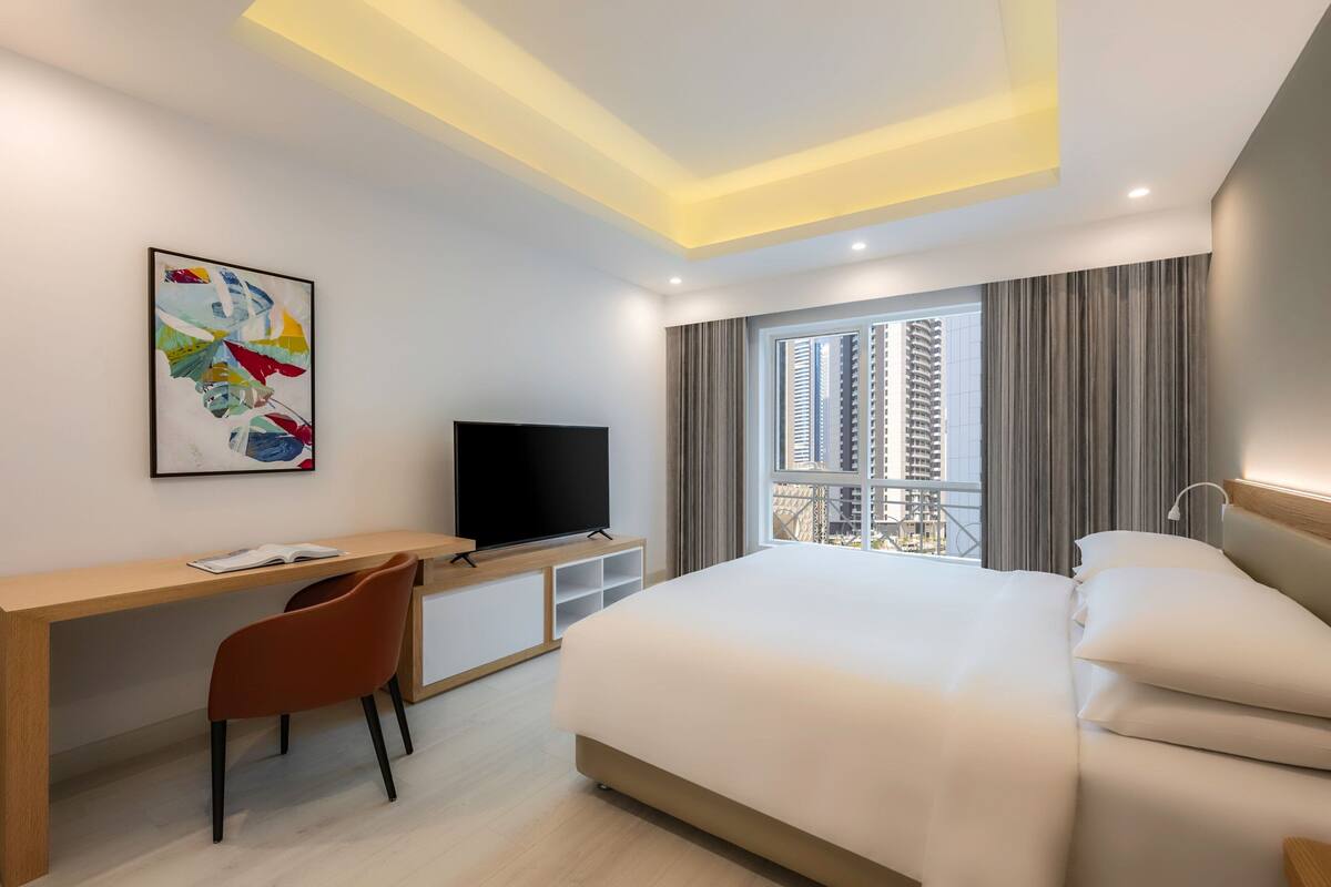 A hotel room with a bed, a desk with a TV, a chair, and a window with curtains.