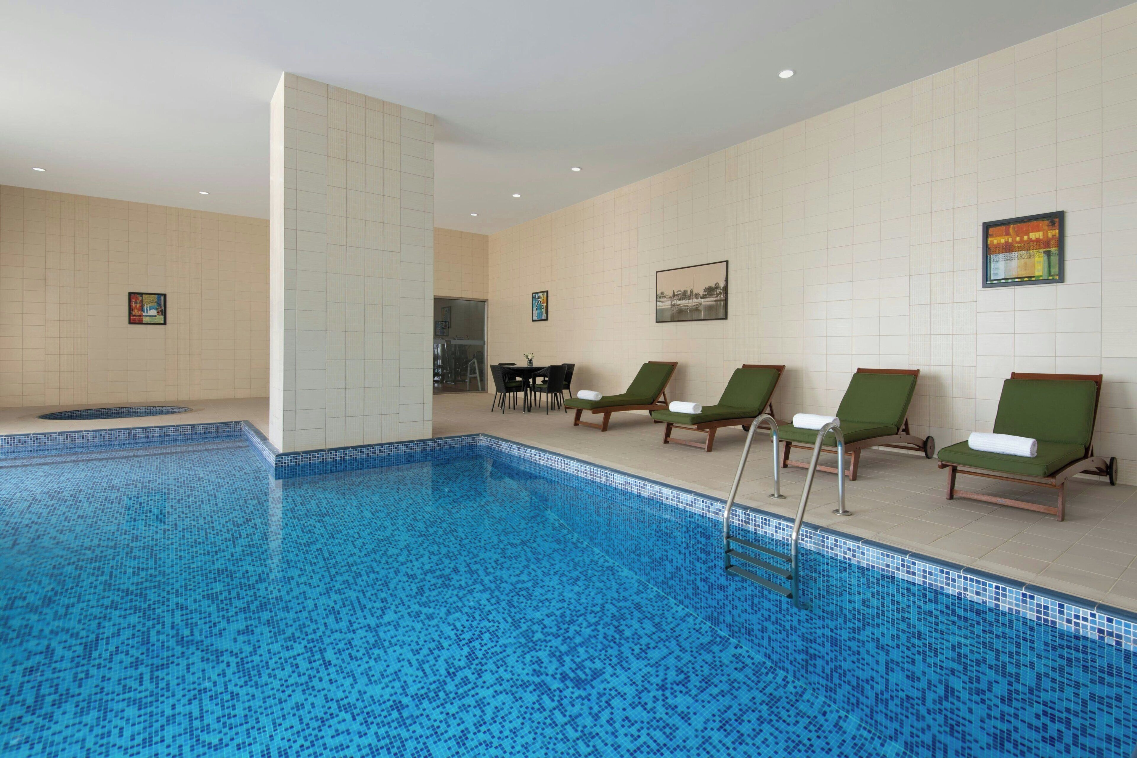 A swimming pool with blue mosaic tiles, surrounded by green lounge chairs with white towels.