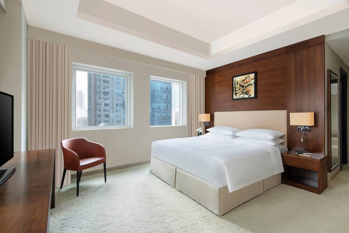 A hotel room with a large bed, two bedside lamps, a TV, a red chair, and a large window with a city view.