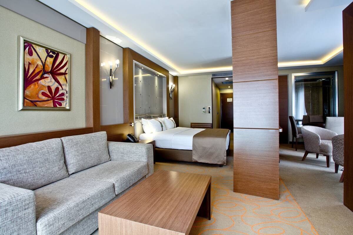 A hotel room with a sofa, a bed, a wooden coffee table, a painting on the wall, and a patterned carpet.