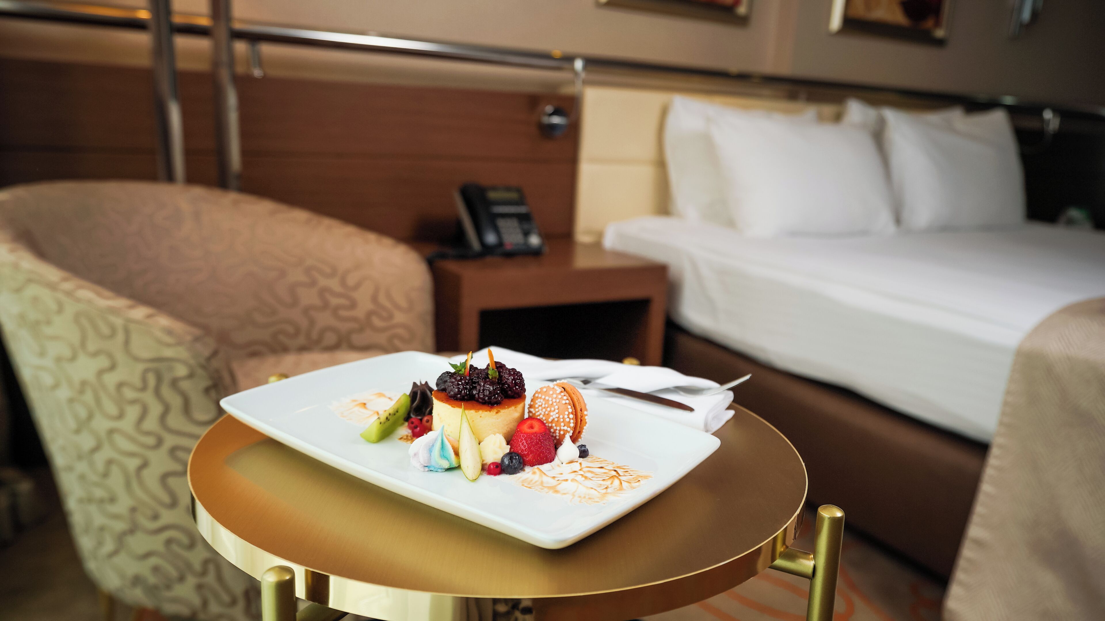 Habitación de hotel con cama, mesita de noche con teléfono y una pequeña mesa redonda con un plato de postre.