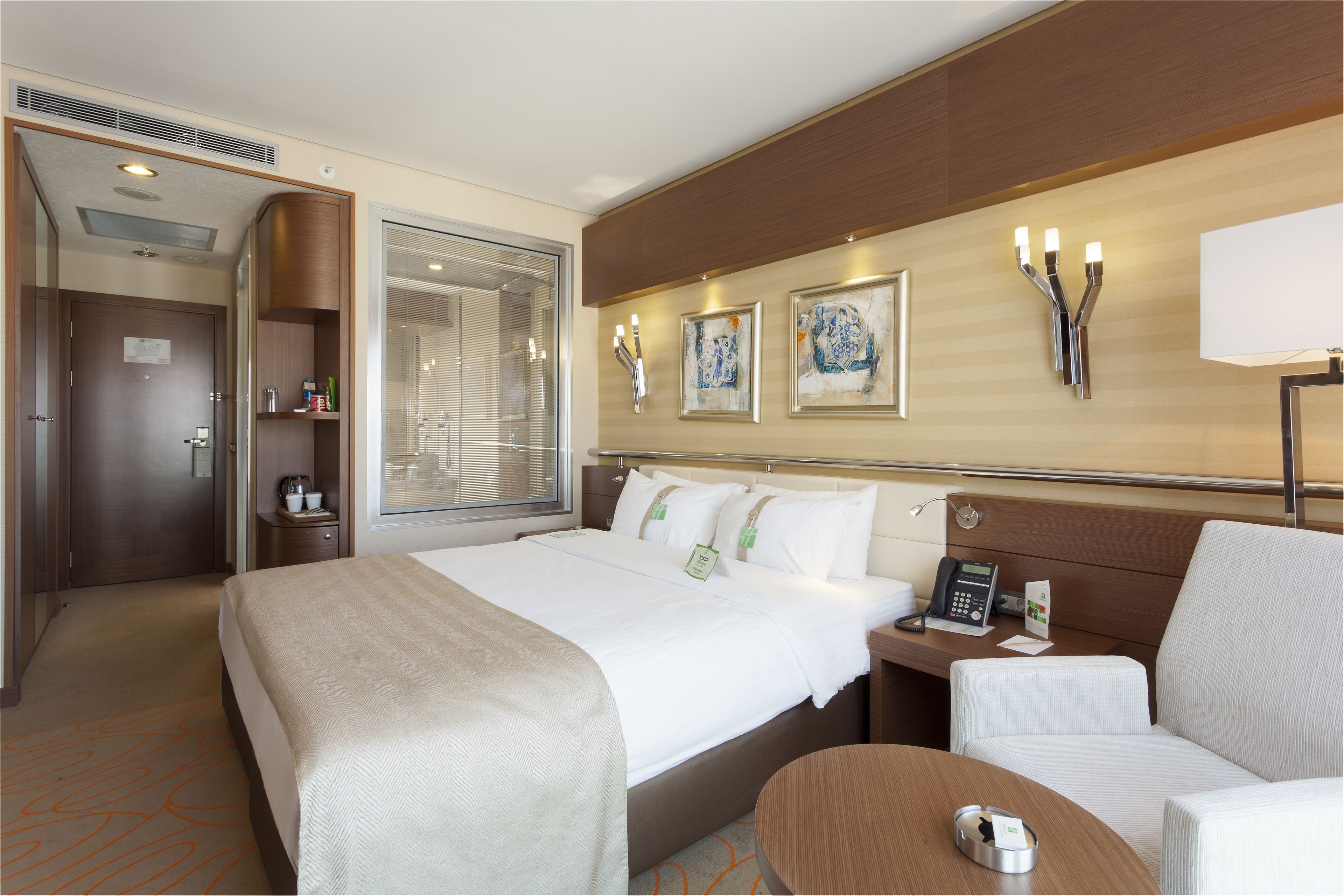 A hotel room with a large bed, a bedside table with a phone and a card, a chair, a small table, and a wall-mounted lamp.