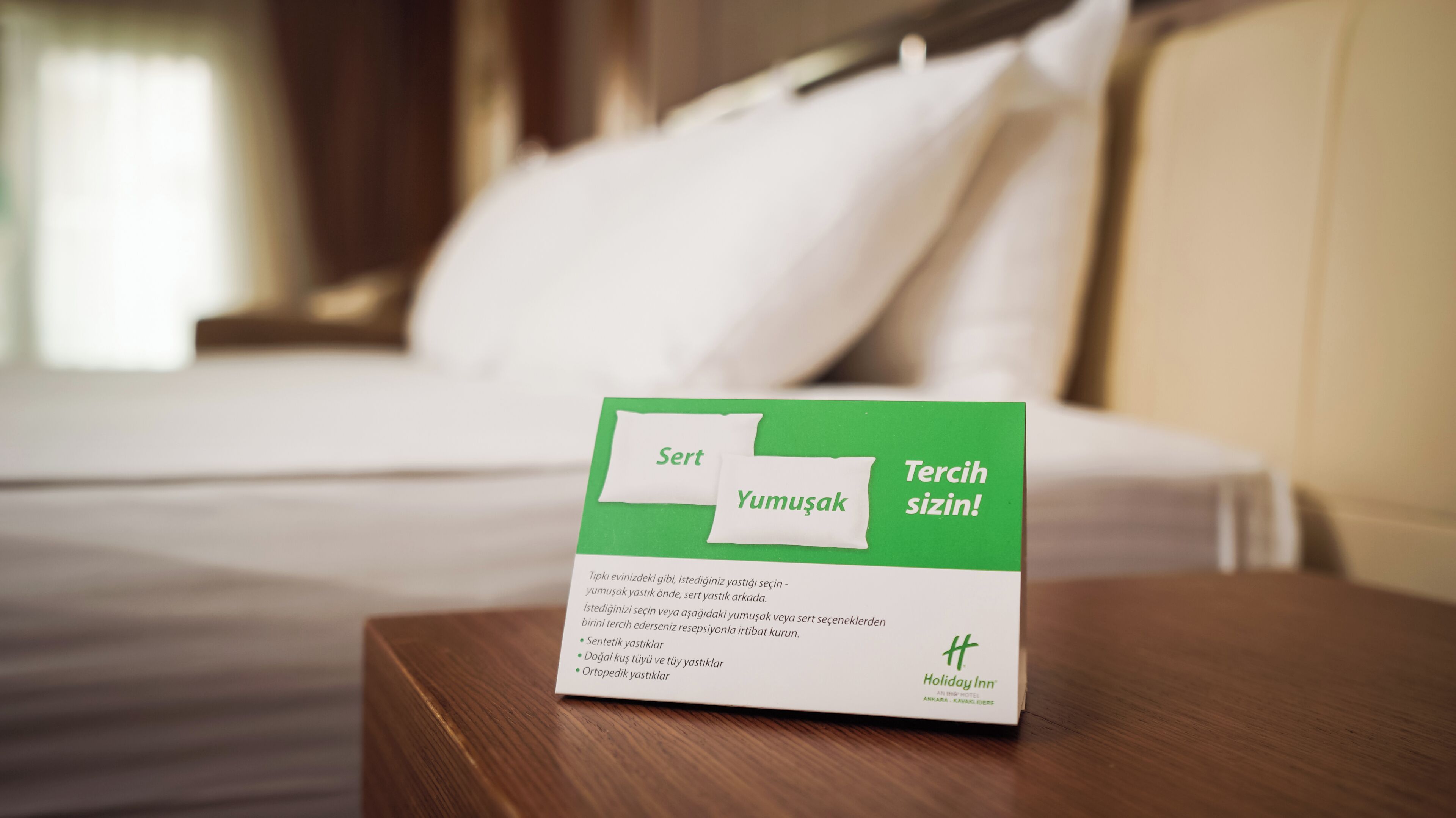 A hotel room with a neatly made bed, a bedside table with a green and white informational card, and a pillow.