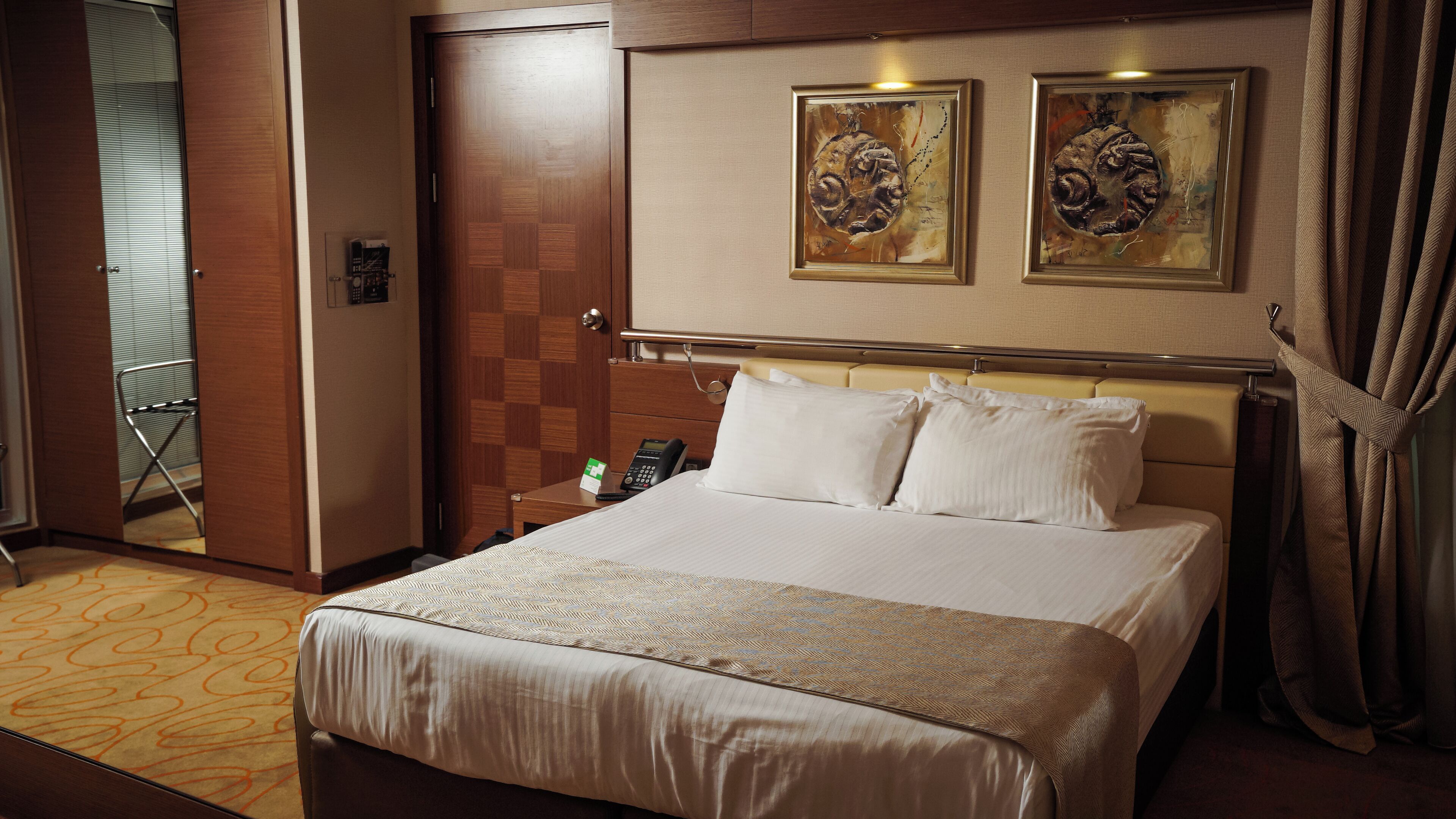 Habitación de hotel con una cama grande, una mesita de noche con un teléfono, dos cuadros enmarcados en la pared y una puerta con mostrador de recepción.