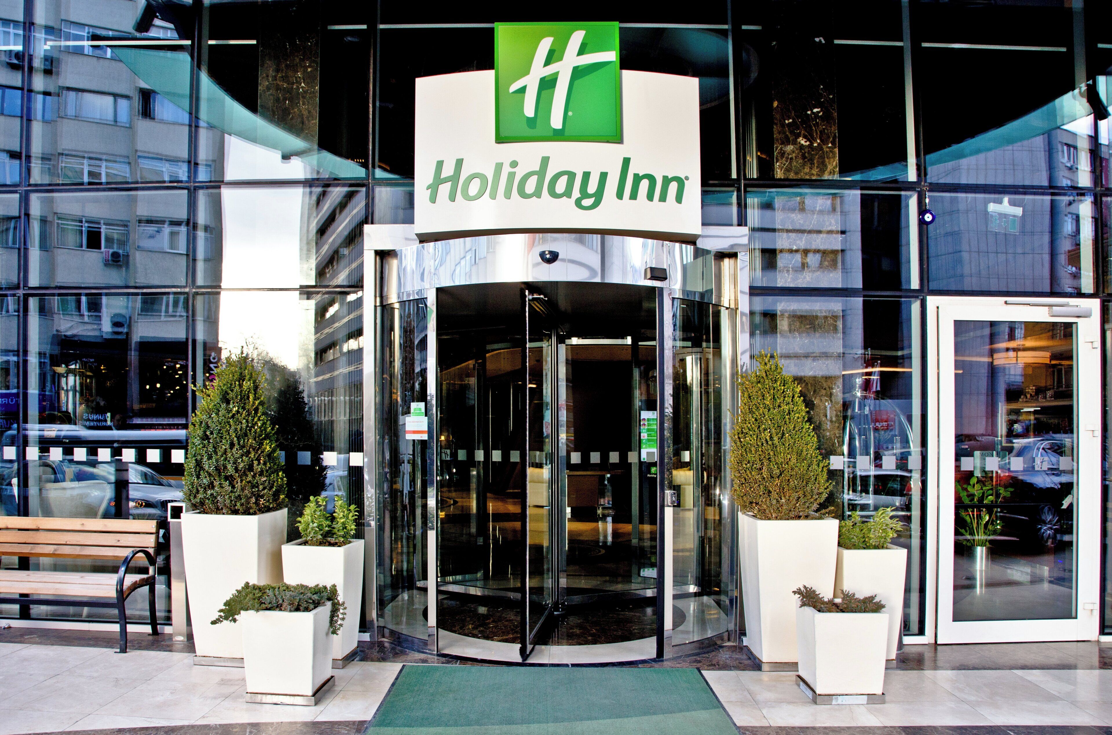 A Holiday Inn hotel entrance with a glass revolving door, potted plants, and a bench outside.