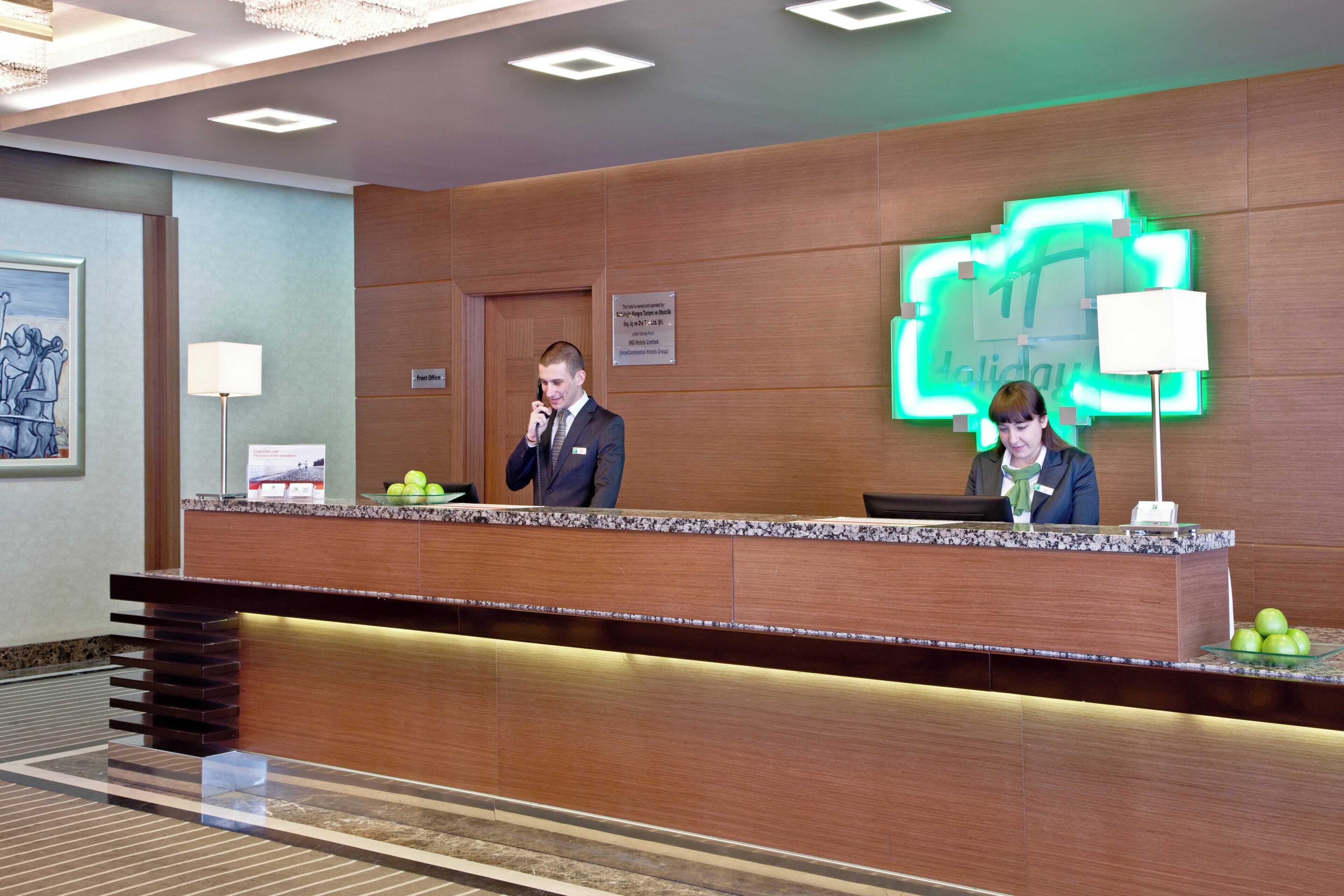 Een hotel receptie met twee medewerkers, een lijst als kunstwerk aan de muur en een neonbord.