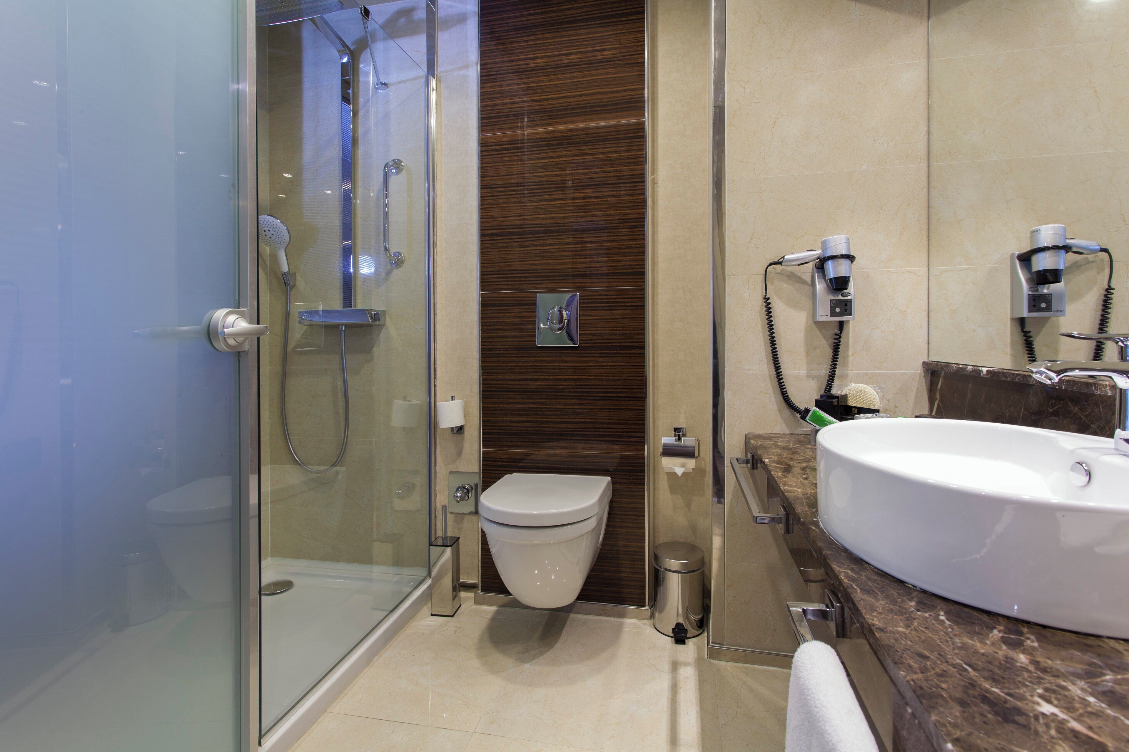 A modern bathroom with a glass-enclosed shower, a toilet, a sink with a marble countertop, and a hairdryer mounted on the wall.