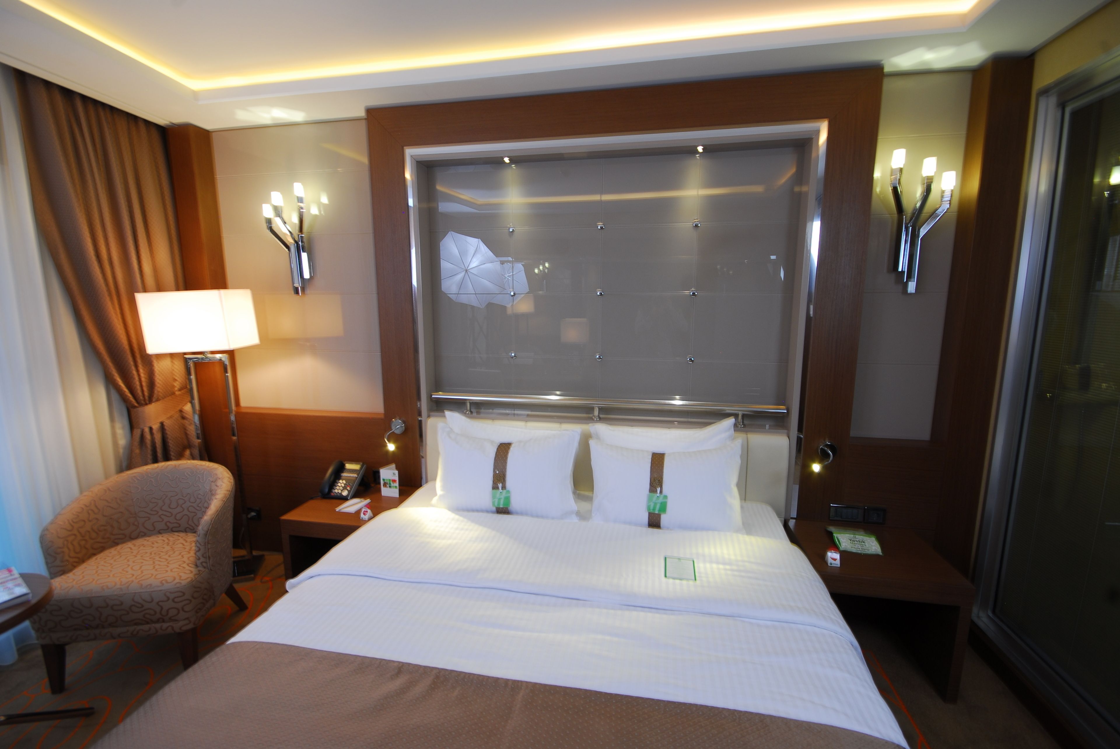 A hotel room with a large bed, a chair, a nightstand with a phone, and a wall-mounted lamp.