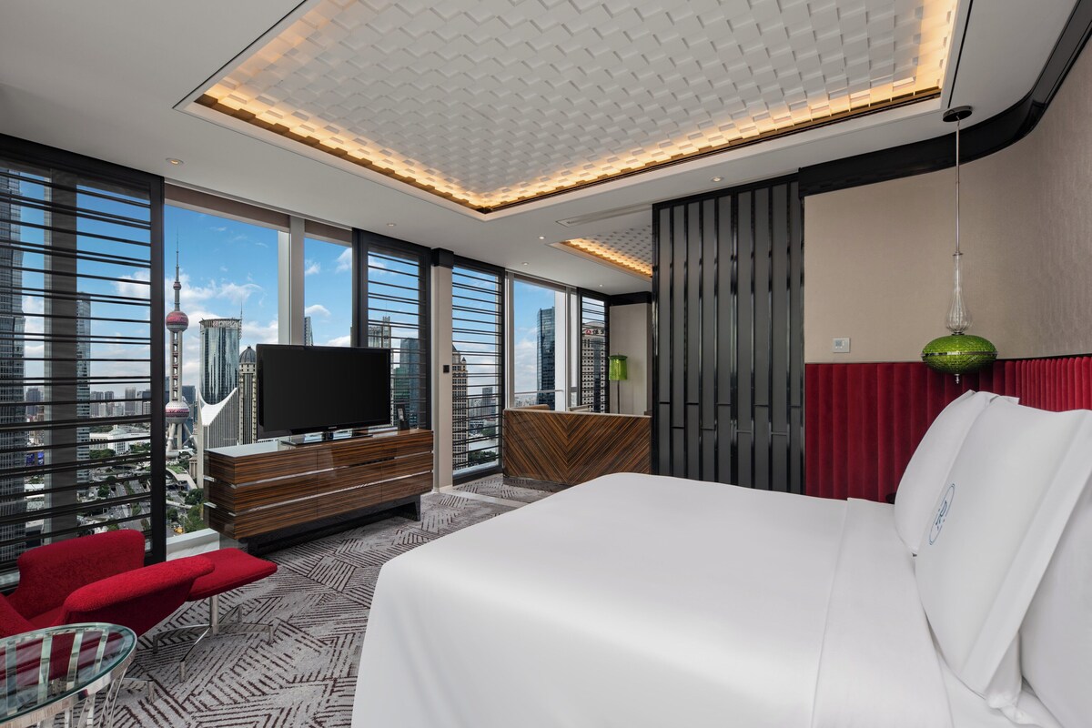 A modern hotel room with a large bed, a TV, and a city view.