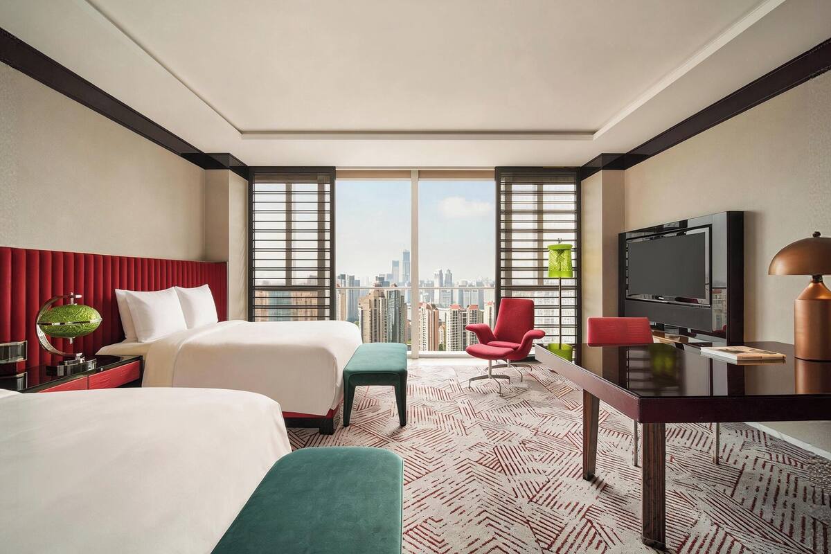 A hotel room with two beds, a desk, a TV, and a city view.