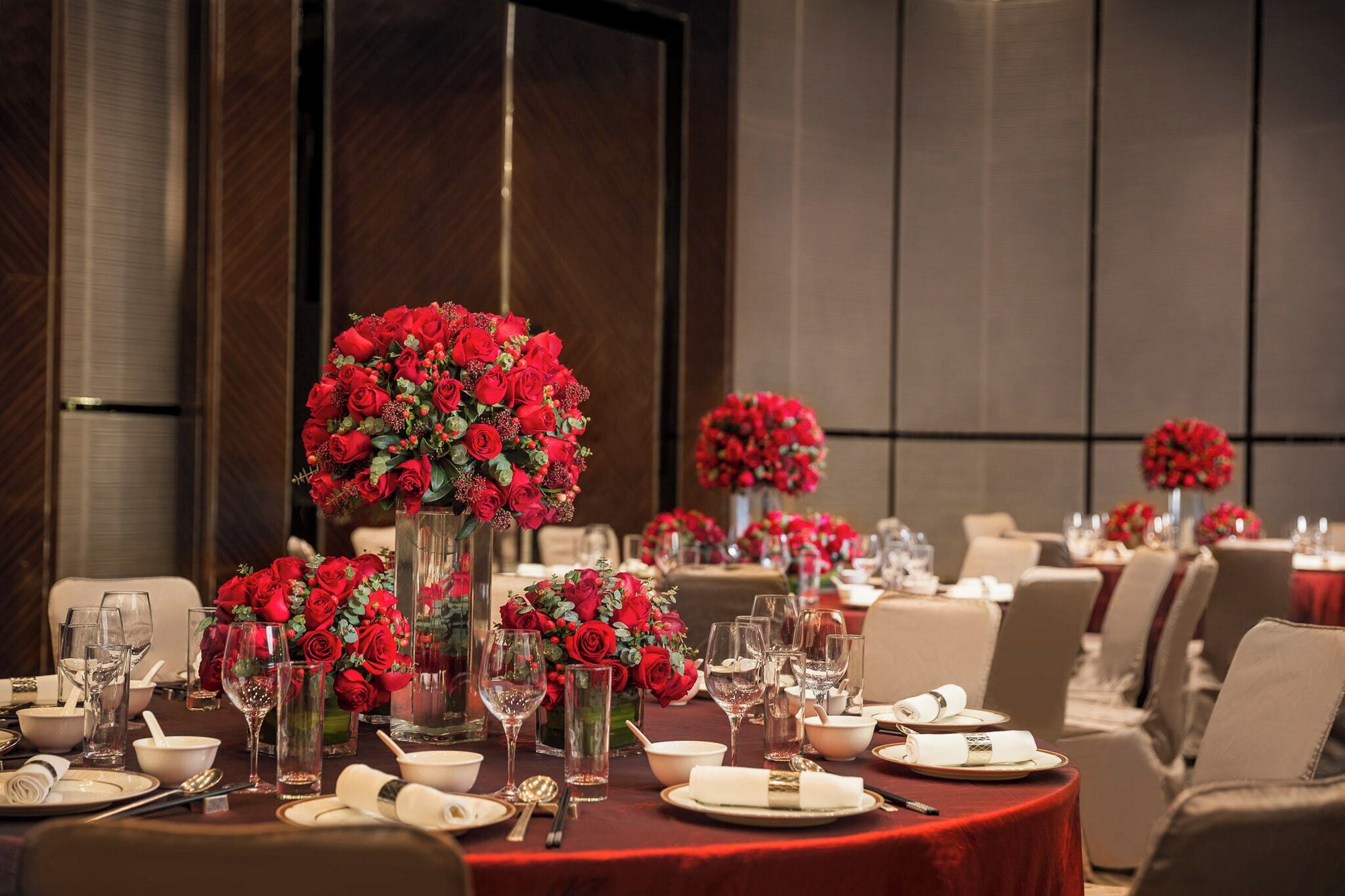 A dining area with round tables set for a meal, each with a centerpiece of red roses in a clear vase.