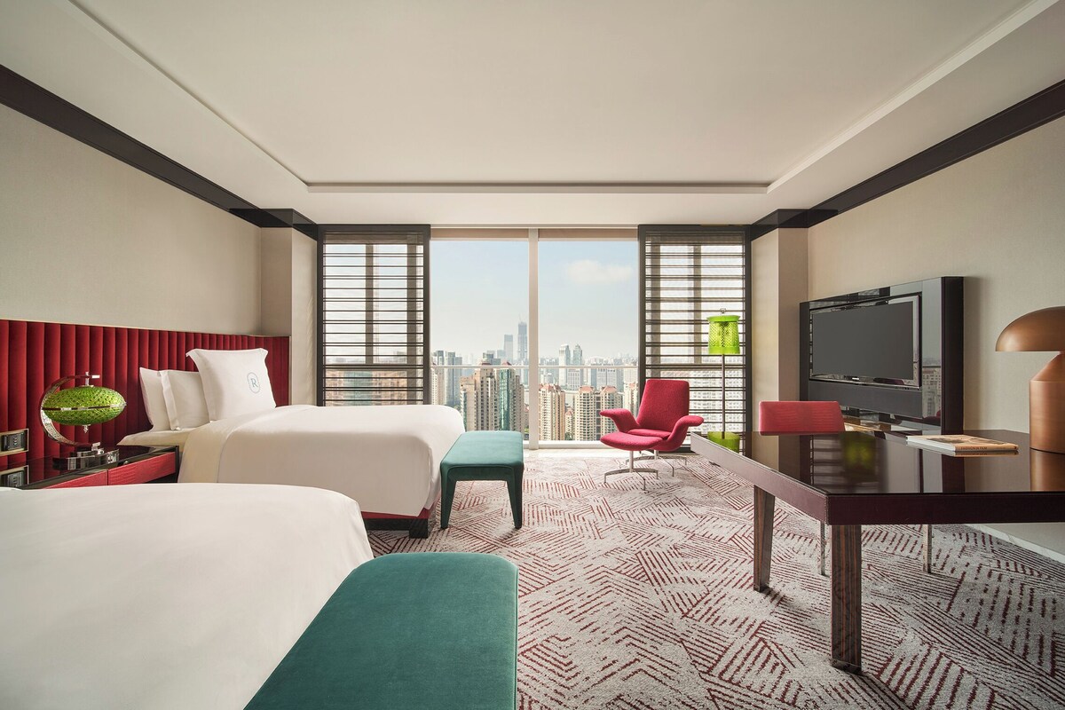 A hotel room with two beds, a desk with a TV, and a city view through large windows.