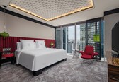 A modern hotel room with a large bed, a red chair, a TV, and a city view.