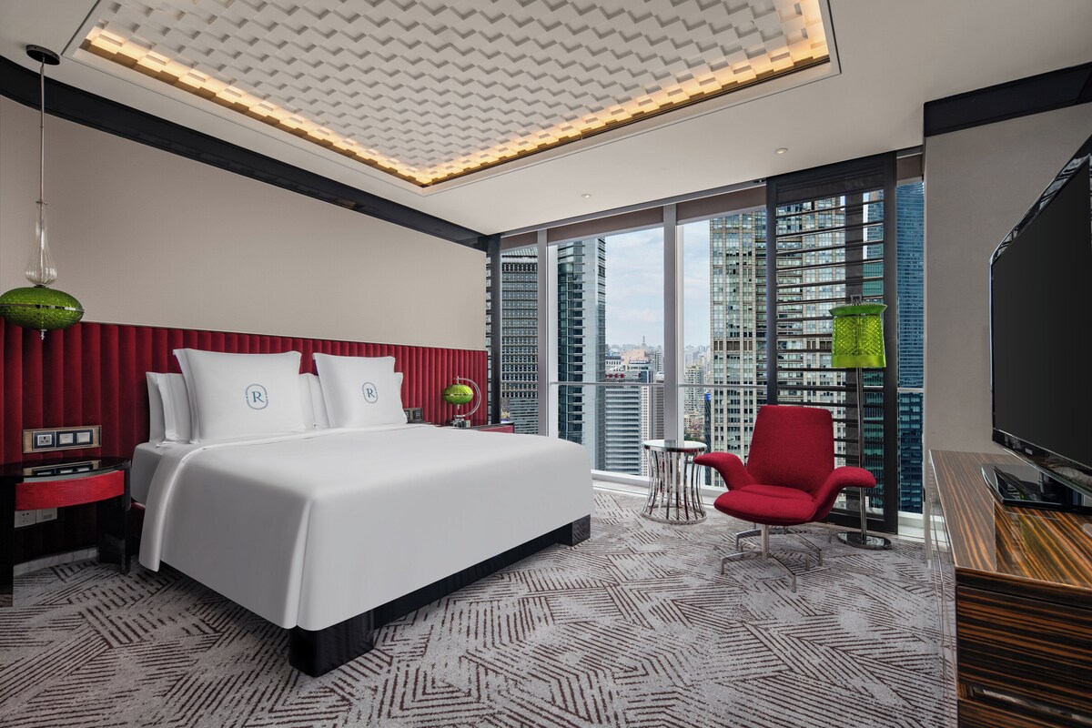 Ein modernes Hotelzimmer mit einem großen Bett, einem roten Sessel, einem Fernseher und Blick auf die Stadt.